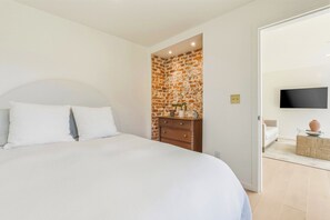 A bedroom with a large bed, a wooden dresser, a brick accent wall, and a flat-screen TV mounted on the wall. - Coastal Sanctuary: Newly Remodeled, Unrivaled Views, Sauna, and Steps to Ferry! (Sausalito)