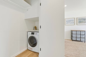 A laundry room with a front-loading washing machine, a shelf with a bottle, and a cart with storage boxes. - Coastal Sanctuary: Newly Remodeled, Unrivaled Views, Sauna, and Steps to Ferry! (Sausalito)