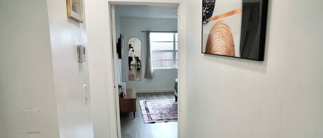 A hallway with a door leading to a room, a mirror, a window with curtains, and a framed artwork on the wall.