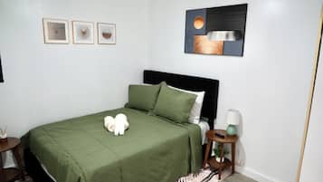 Una cama bien hecha con colcha verde, cabecera y dos almohadas. En la pared, hay tres cuadros enmarcados, una mesita redonda con lámpara y una mesita de noche de madera con una planta.