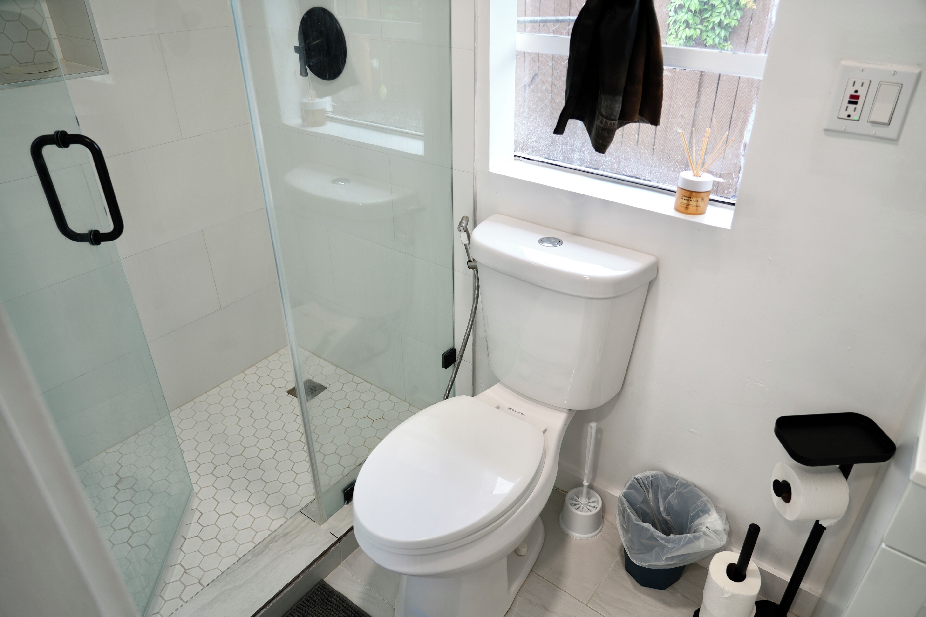 Un baño con inodoro, cabina de ducha de vidrio, ventana con vistas y un estante con artículos de aseo.
