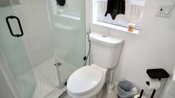 Un baño con inodoro, cabina de ducha de vidrio, ventana con vistas y un estante con artículos de aseo.