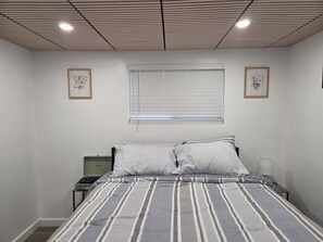 A bedroom with a bed, a bedside table, a window with blinds, and two framed pictures on the wall. - Centrally located 1-bedroom apartment close to downtown Wifi AC washer/dryer (Twin Falls)