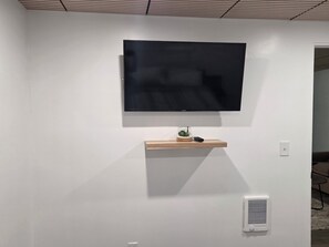 A flat-screen TV mounted on a white wall, a small wooden shelf with a plant and a remote, and a white vent. - Centrally located 1-bedroom apartment close to downtown Wifi AC washer/dryer (Twin Falls)