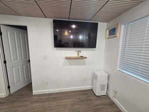 A room with a white wall, a mounted TV, a small wooden shelf with a vase, a white air conditioner, and a door leading to another room. - Centrally located 1-bedroom apartment close to downtown Wifi AC washer/dryer (Twin Falls)