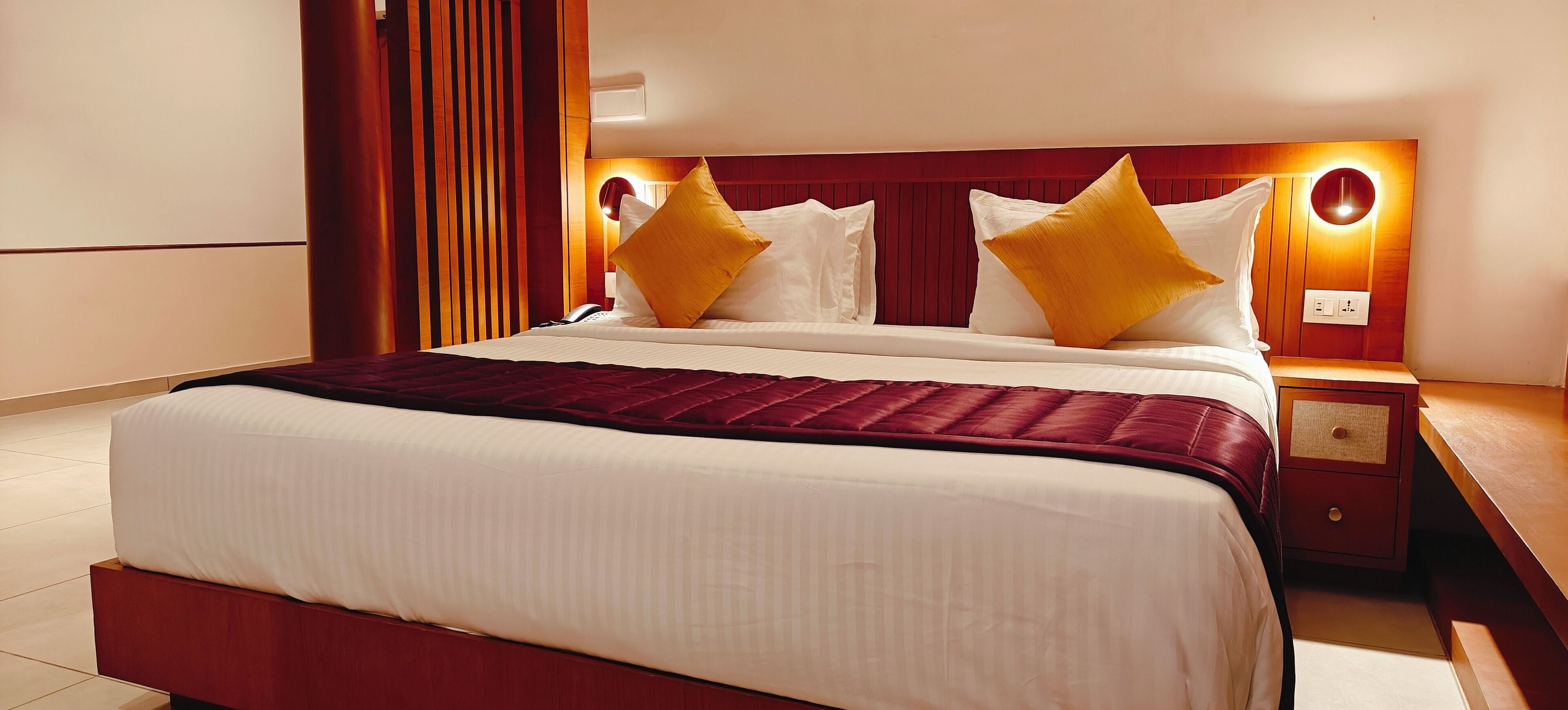 A hotel room with a large bed, a bedside table, a nightstand, and a wall-mounted lamp.