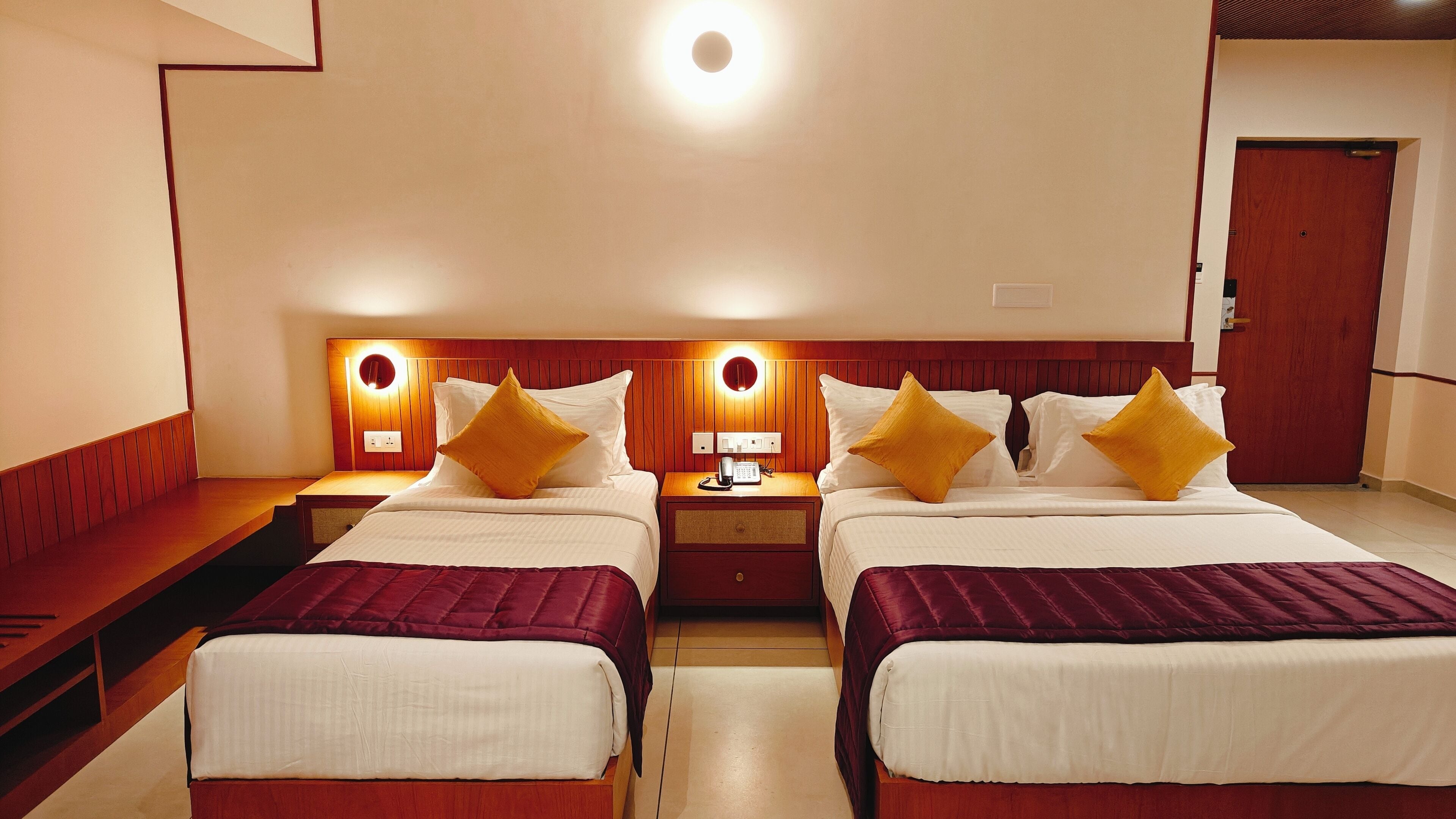 A hotel room with two beds, a bedside table with a lamp, a phone, and a wall-mounted light fixture.