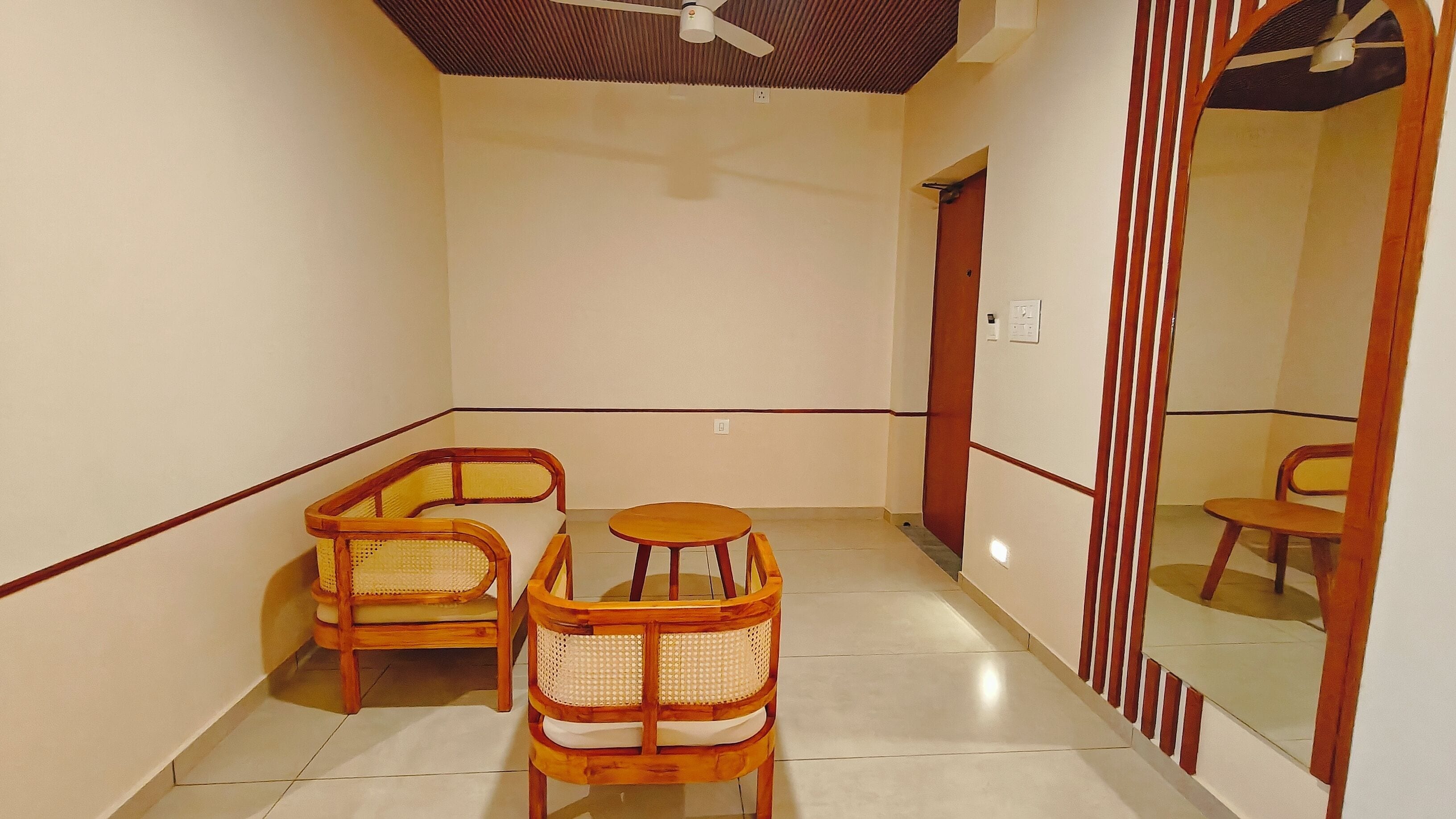Una habitación con dos sillones de mimbre, una mesa redonda pequeña, un ventilador de techo y un espejo que refleja la habitación.