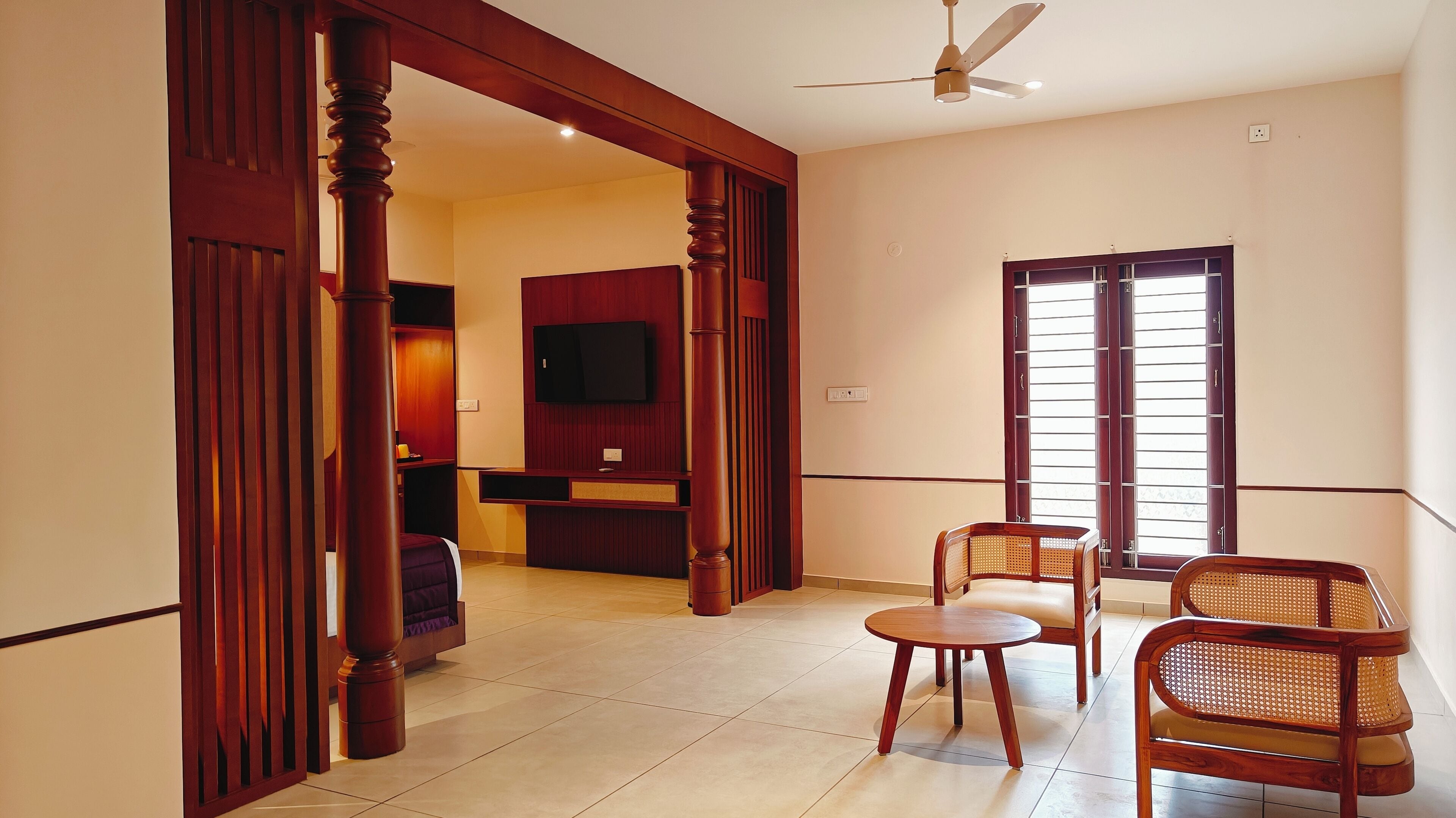 A hotel room with a wooden floor, a ceiling fan, a television mounted on the wall, a window with blinds, and a seating area with two wicker chairs and a small table.