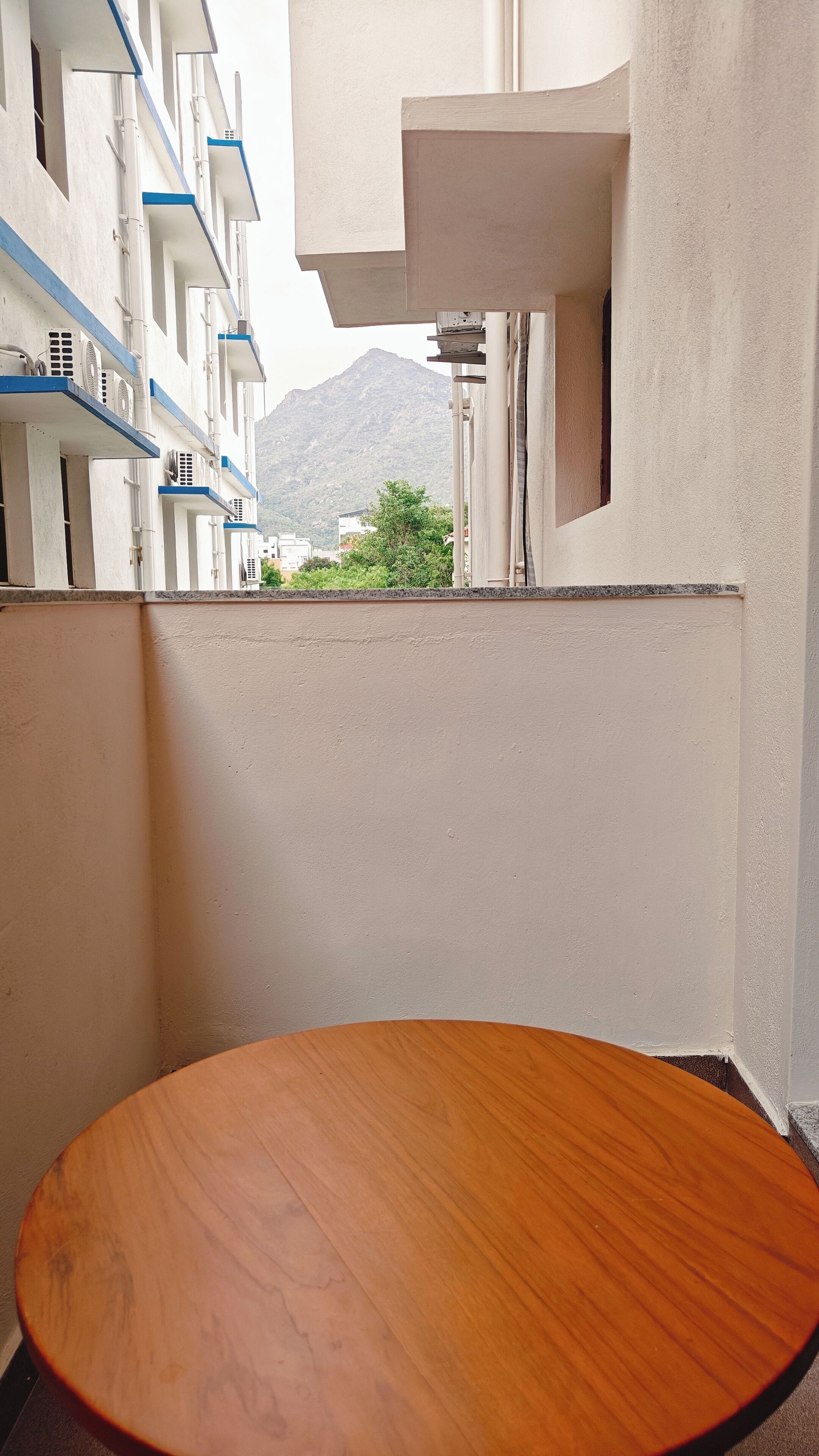 Rincón de una habitación de hotel con una mesa de madera, vista a una montaña y un edificio con detalles azules.