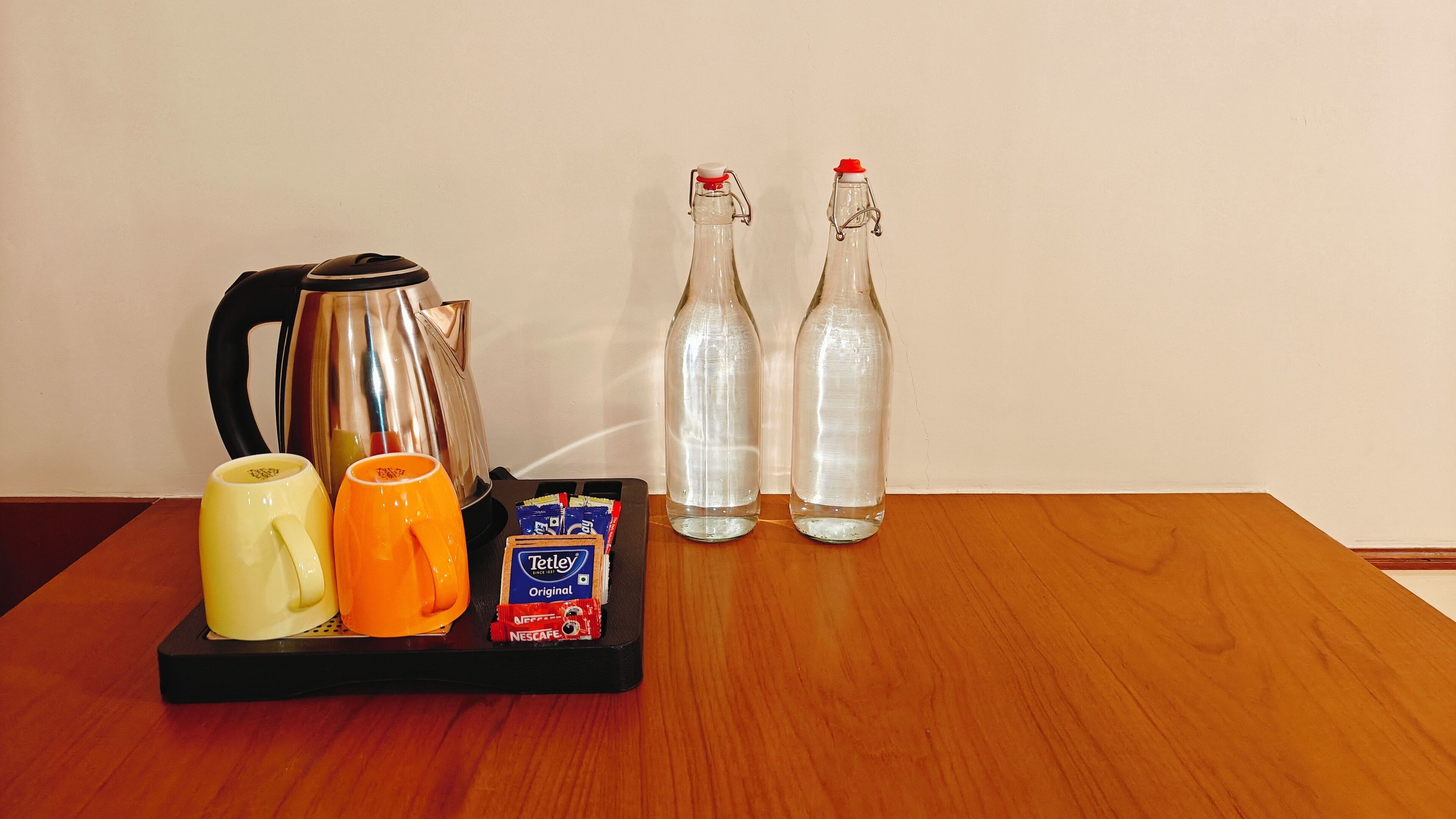A tray with a kettle, two bottles, a yellow mug, an orange mug, and two packets of coffee on a wooden table.