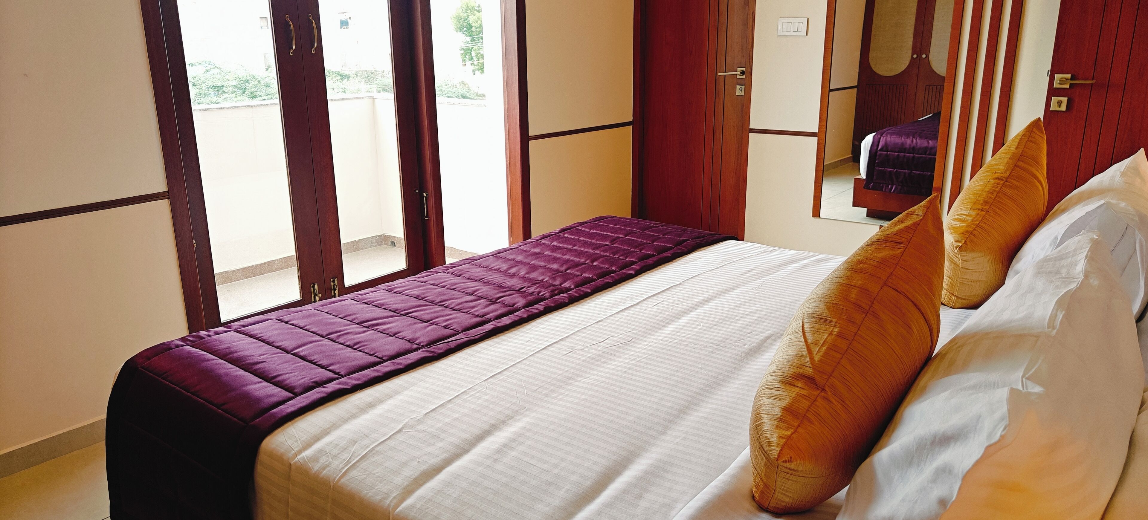 Una cama con colcha morada y ropa de cama blanca, cabecera de madera y vista al exterior por una ventana.