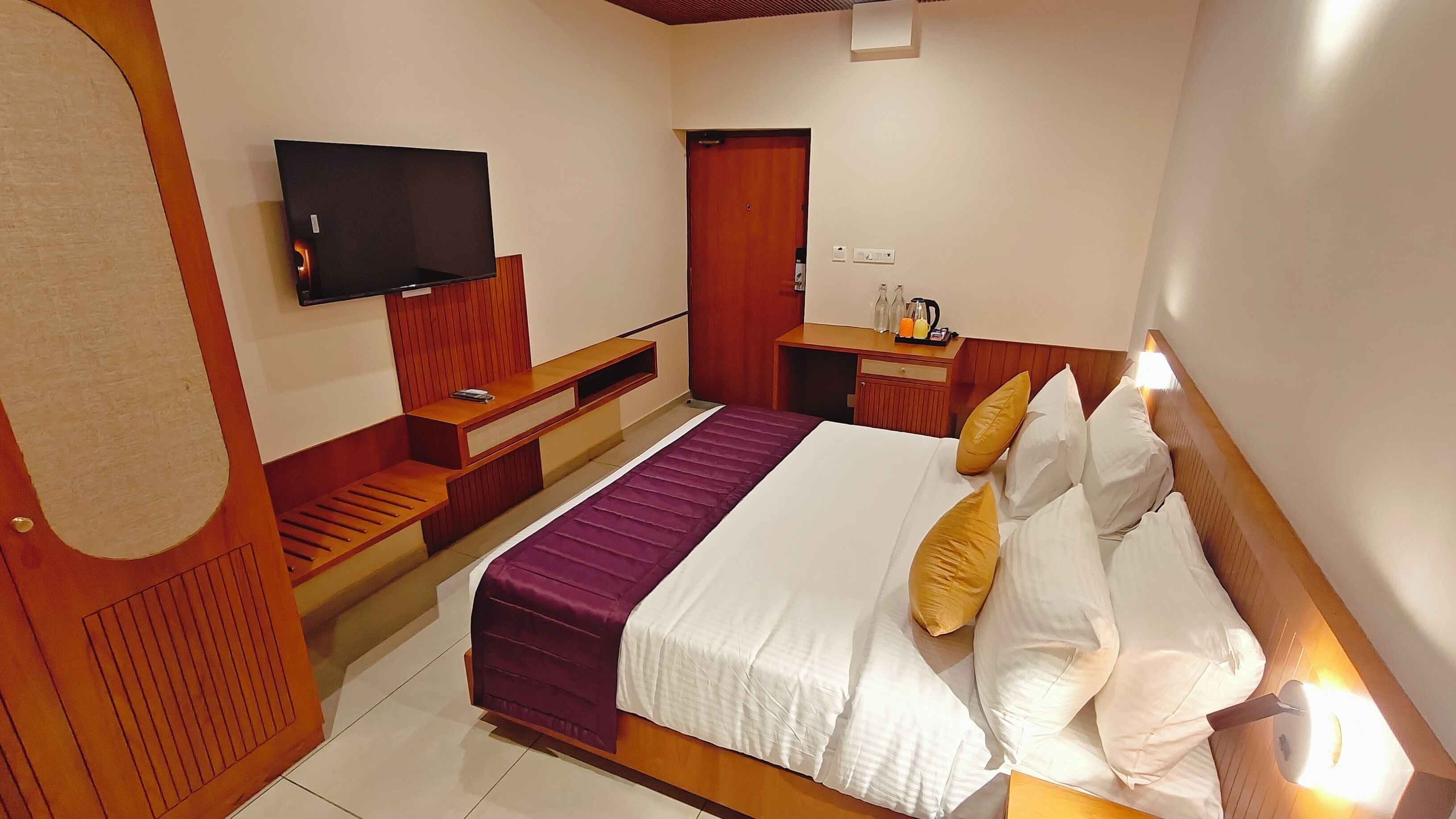 Habitación de hotel con una cama, un televisor montado en la pared, un banco de madera, una mesita de noche y una puerta.