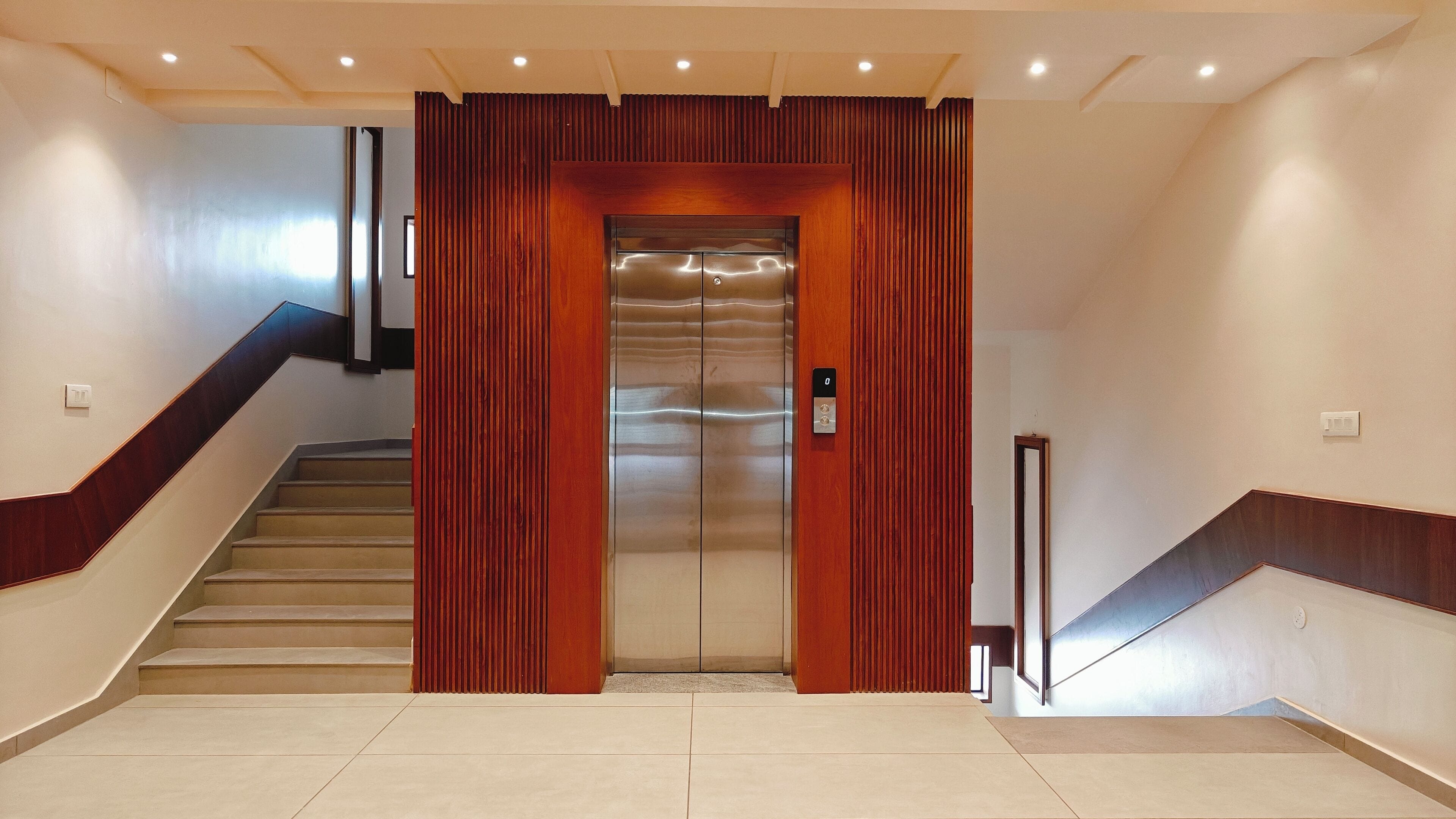 Un ascenseur moderne aux finitions métalliques, encadré de panneaux en bois et agrémenté d’un escalier doté d’une rampe en bois.