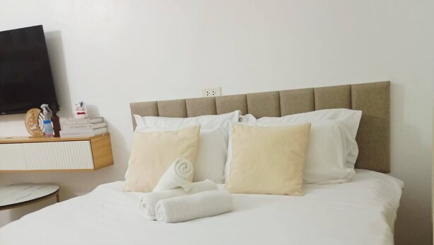 Una cama bien hecha con ropa blanca y cabecera, una mesita de noche con un libro y una botella, y un televisor montado en la pared.