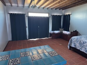 Een slaapkamer met een bed, een bankje, een raam met gordijnen en een betegelde vloer.
