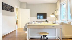 Ein modernes Wohnzimmer mit einem weißen Schreibtisch in der Mitte, einer gemütlichen Couch, einem an der Wand befestigten Fernseher und einem Fenster mit Vorhängen.