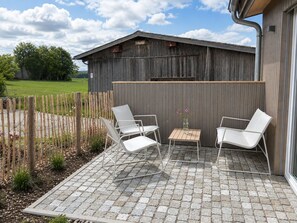 Eine Terrasse mit zwei weißen Stühlen und einem kleinen Tisch, ein Holzzaun und ein Gebäude im Hintergrund.