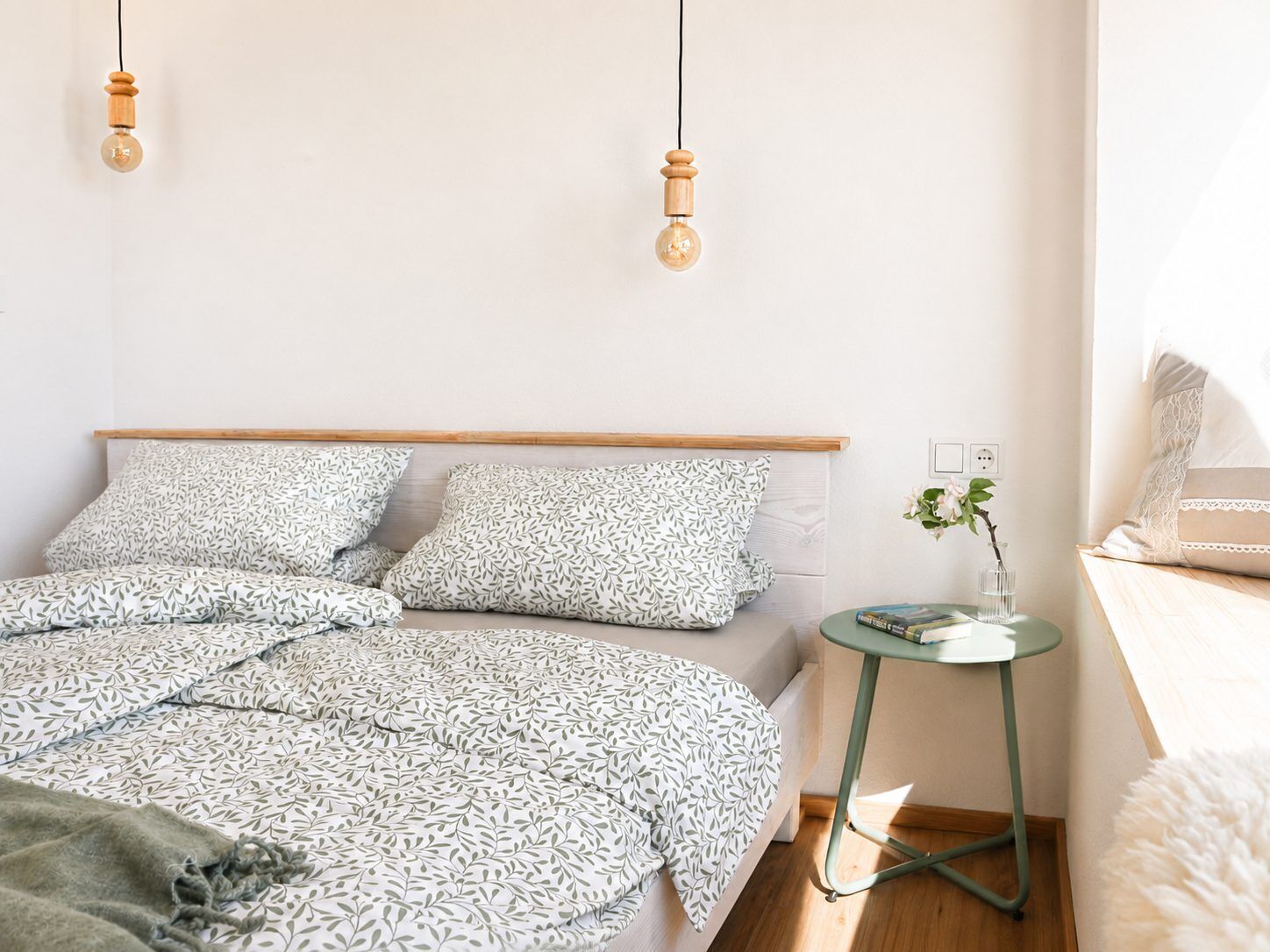 A bedroom with a bed, pillows, a bedside table, a lamp, and a wall socket.