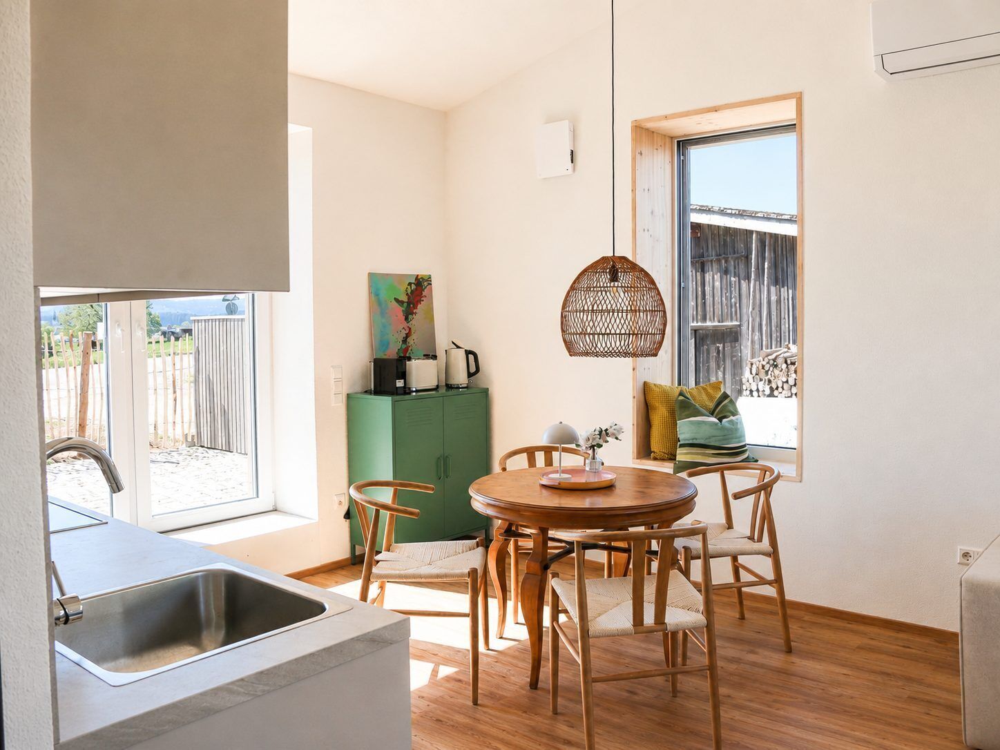A compact kitchen with a sink, a dining area with a round table and chairs, a green cabinet, and a window with a view of the outside.