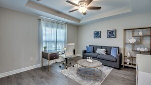 A modern living room with a ceiling fan, a grey sofa with blue cushions, a round coffee table, and a desk with a chair. - Luxury Cape Coral Oasis: Pool, Sleeps 8 (Cape Coral)