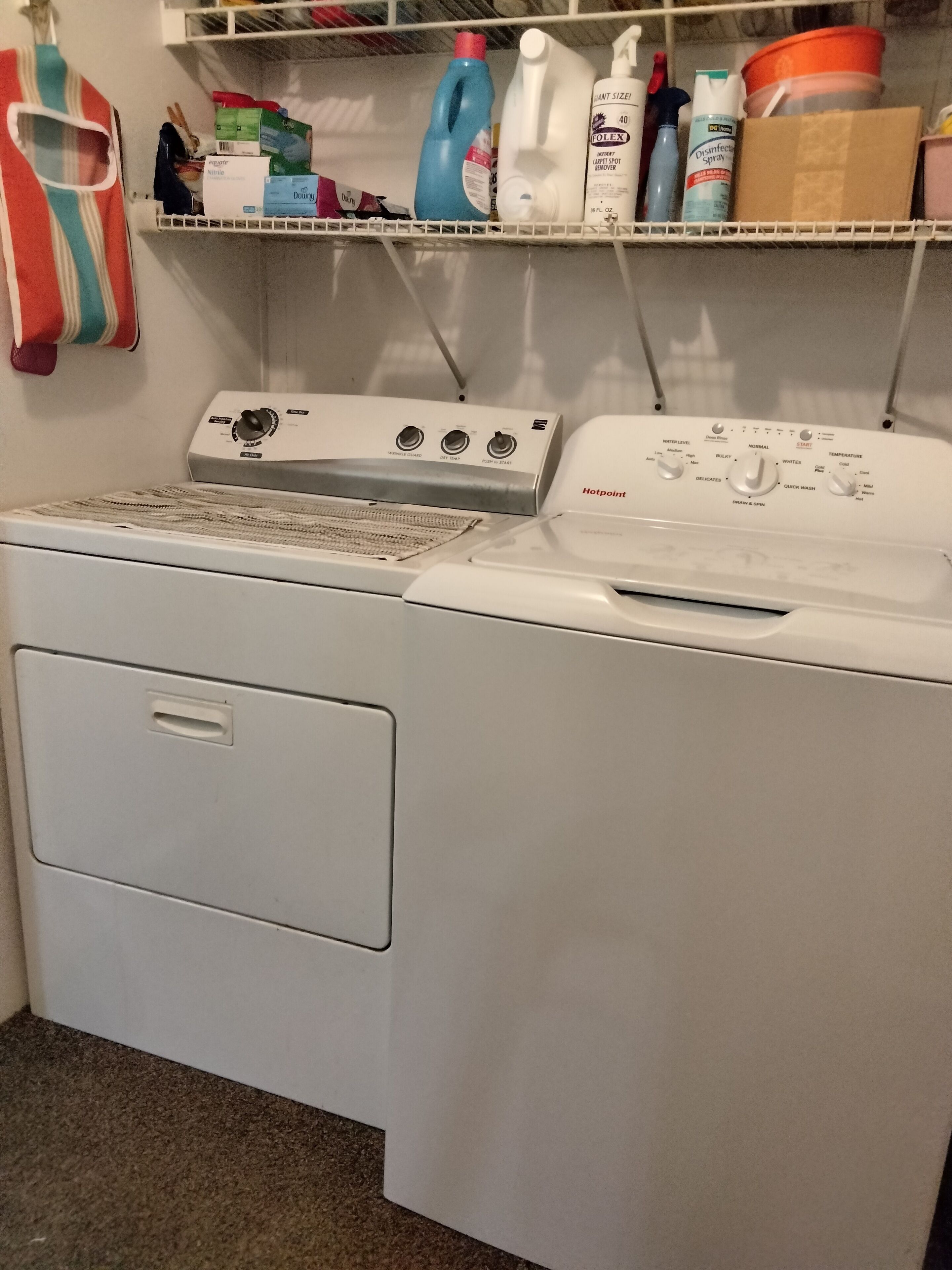 A front-loading washing machine with a control panel, a dryer, and a shelf above containing cleaning supplies.
