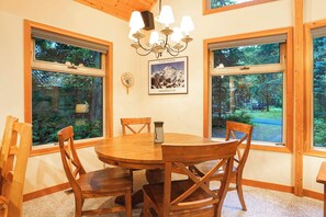 A dining area with a wooden table and chairs, large windows with a view of trees, and a chandelier. - Wildwood Snowline Cabin Near Mt Baker (Deming)