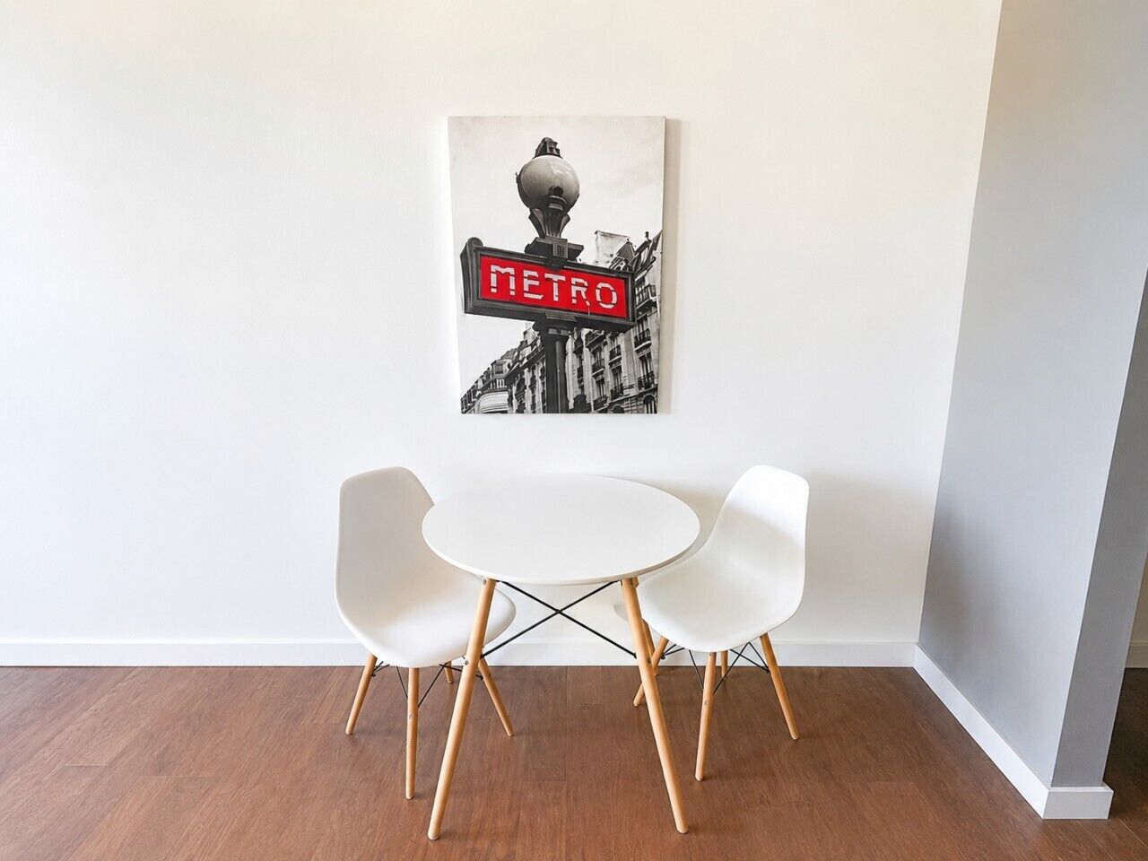 A room with a white table and two white chairs, a wall-mounted picture, and a wooden floor.