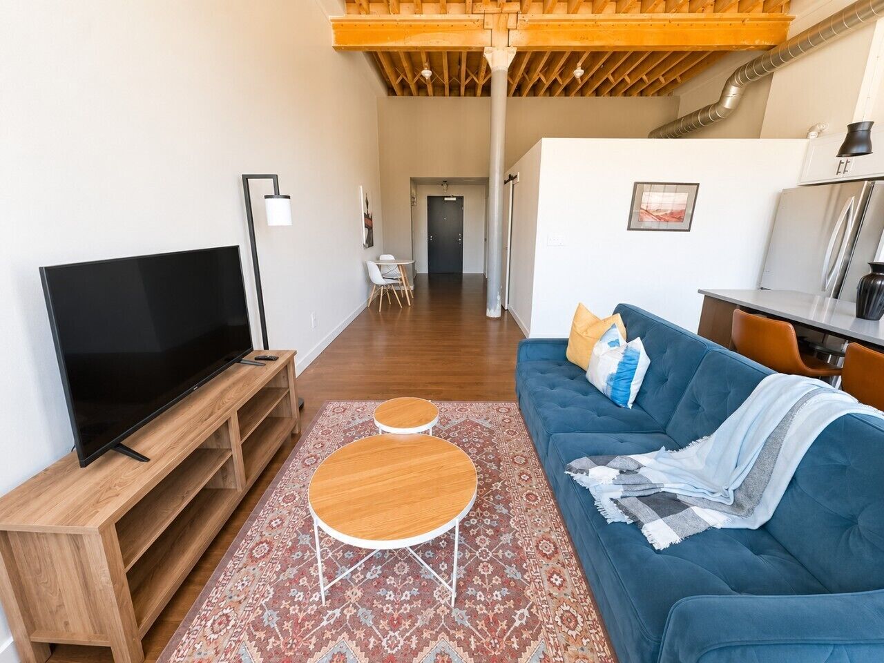 Una sala de estar moderna con un televisor de pantalla plana, un sofá azul, dos mesas redondas de madera y una alfombra estampada.