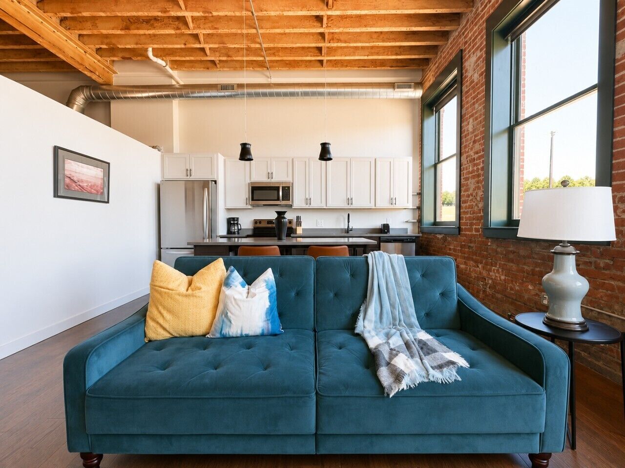 Ein modernes Wohnzimmer mit einem türkisblauen Sofa, einem Couchtisch und einer Lampe. Im Hintergrund ist eine Küchenzeile mit weißen Schränken und einer Mikrowelle zu sehen.