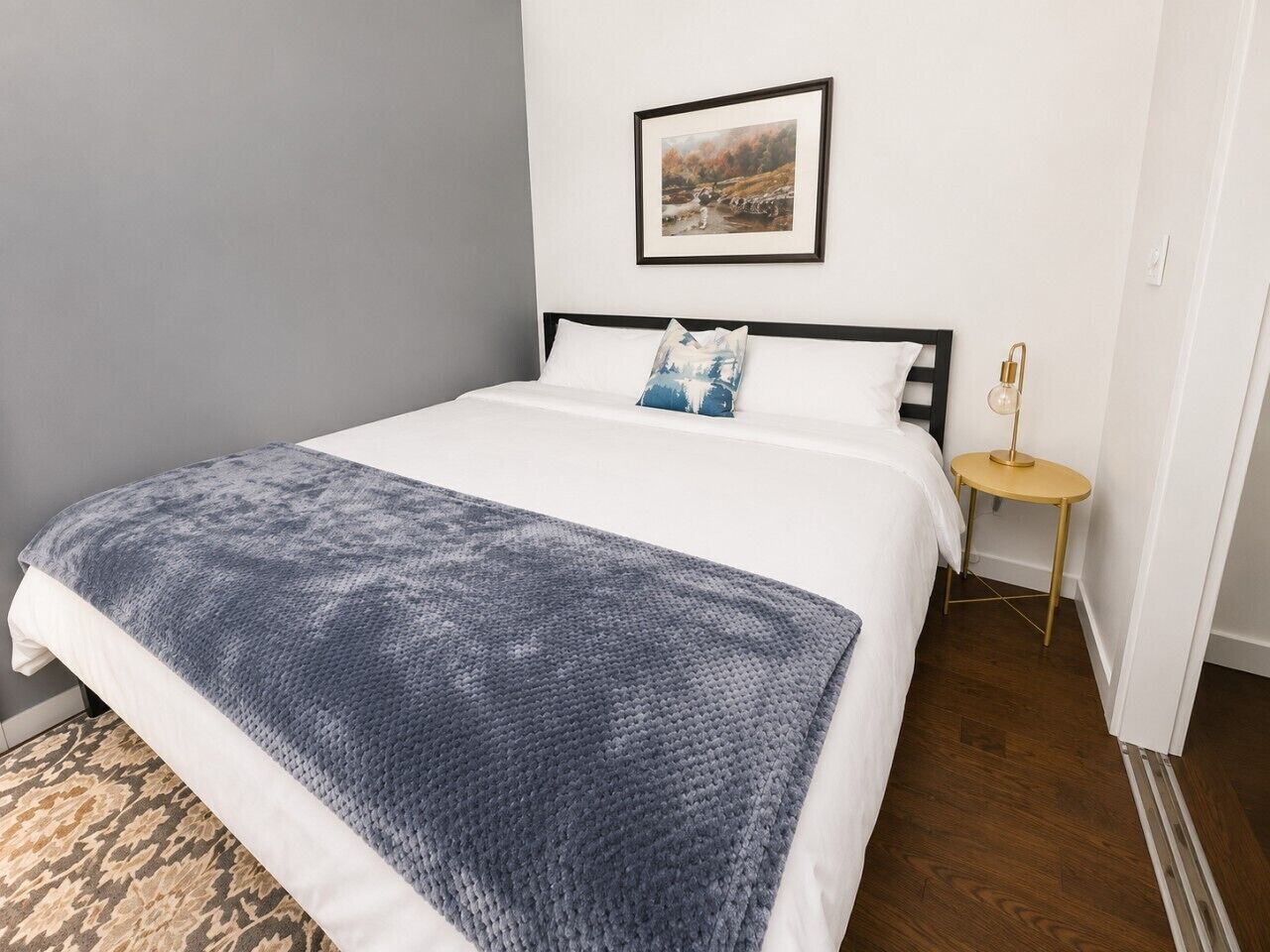 A neatly made bed with a white and blue blanket, a small round table with a lamp, and a framed picture on the wall.