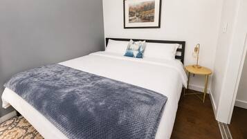 A neatly made bed with a white and blue blanket, a small round table with a lamp, and a framed picture on the wall.
