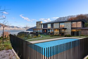 Una casa moderna de dos pisos con piscina, rodeada por una cerca negra y con montañas al fondo.