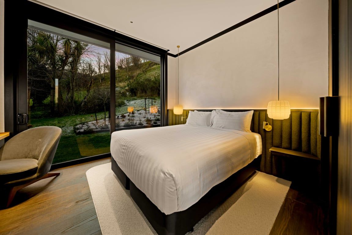Um quarto moderno com uma cama grande, uma cadeira, vista para o exterior por uma porta deslizante e uma mesa de cabeceira com abajur.