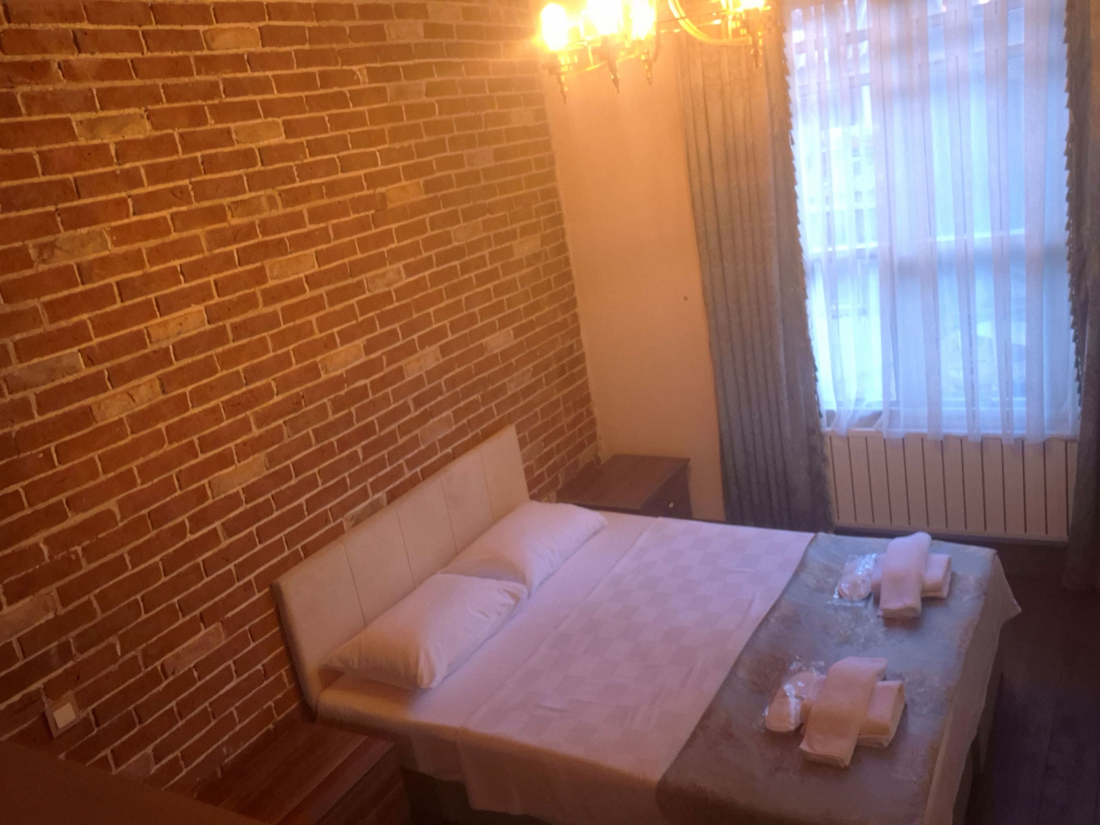 A hotel room with a brick wall, a bed with white bedding, a nightstand, a table with towels, and a window with curtains.