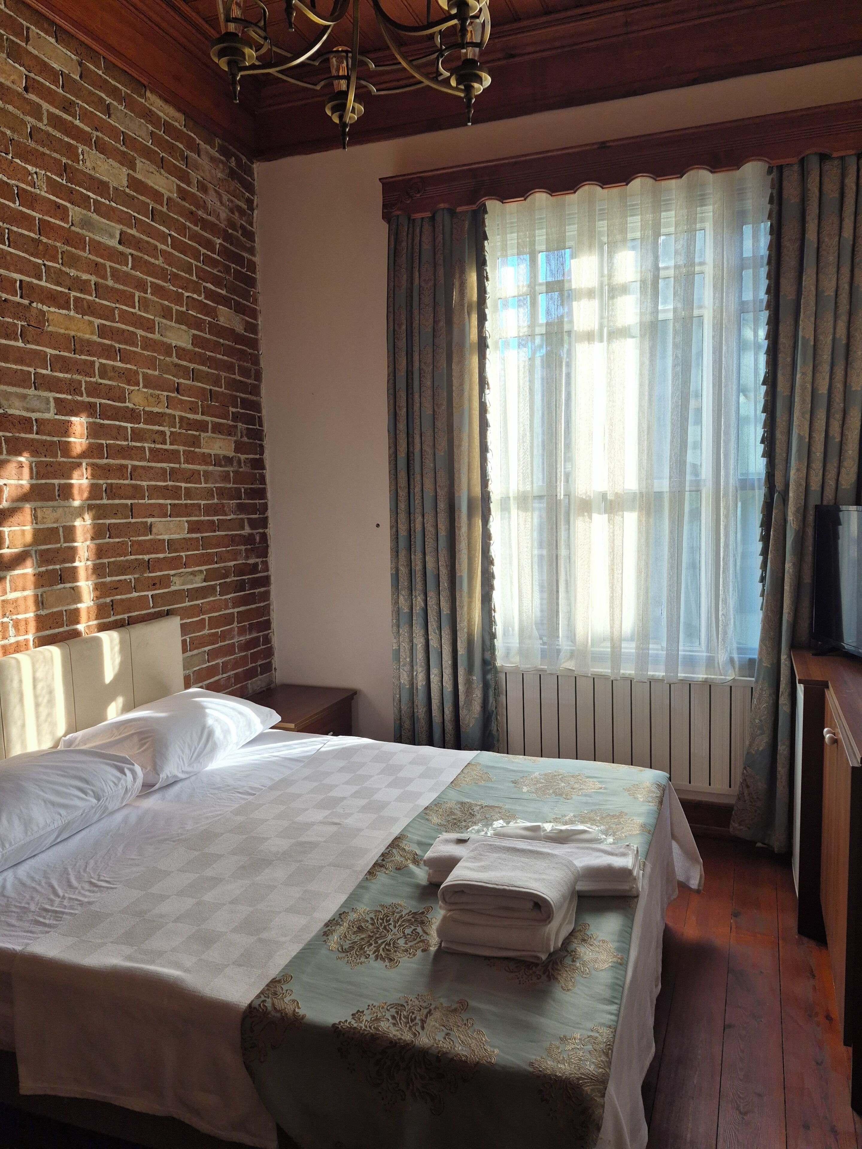 A hotel room with a brick wall, a bed with a checkered bedspread, a nightstand, a desk with a television, and a window with sheer curtains.