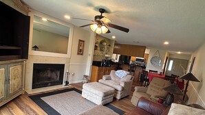 A living room with a fireplace, a ceiling fan, and a kitchen in the background. - Gorgeous 2-bedroom condo with beautiful pool, hot tub, and more in Hot Springs (Hot Springs)