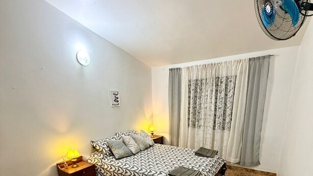 Un dormitorio con cama, mesitas de noche, un ventilador de pared y una ventana con cortinas.