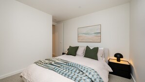 A bedroom with a bed, pillows, a blanket, a nightstand, a lamp, and a painting on the wall. - Dromana Beach Suites - 104 (Dromana)