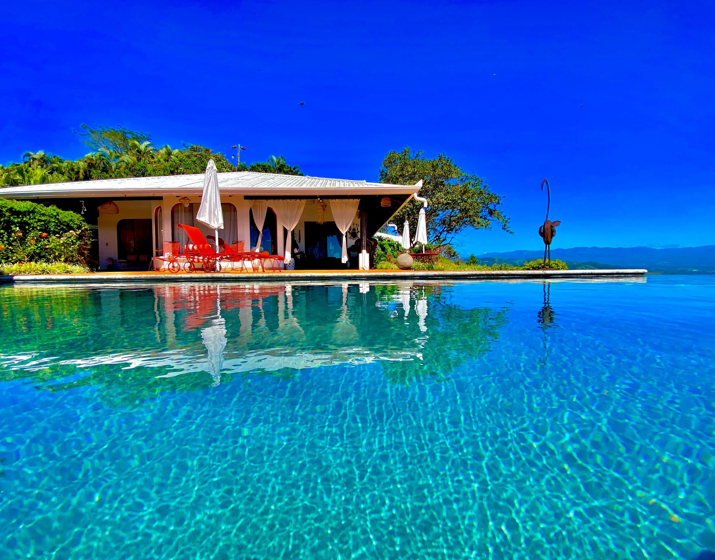 Una piscina cristalina con un área de descanso, sillones rojos y un espacio cubierto para sentarse.