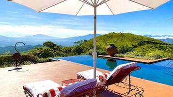 Área da piscina com espreguiçadeiras, um guarda-sol e vista para as montanhas.