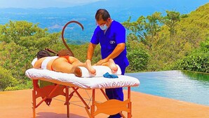 Ein Massagetisch im Freien mit einer Person, die eine Massage erhält, umgeben von Grünpflanzen und Blick auf den Ozean.
