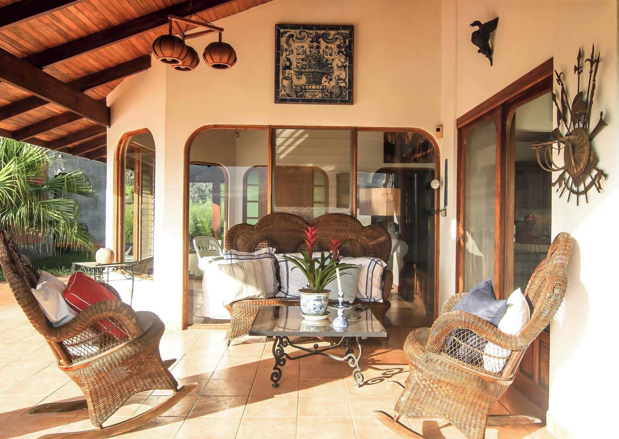 A covered patio area with wicker furniture, a central table with a vase, and a decorative wall piece.