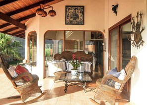 Uma área de varanda coberta com móveis de vime, uma mesa central com um vaso e um elemento decorativo na parede.