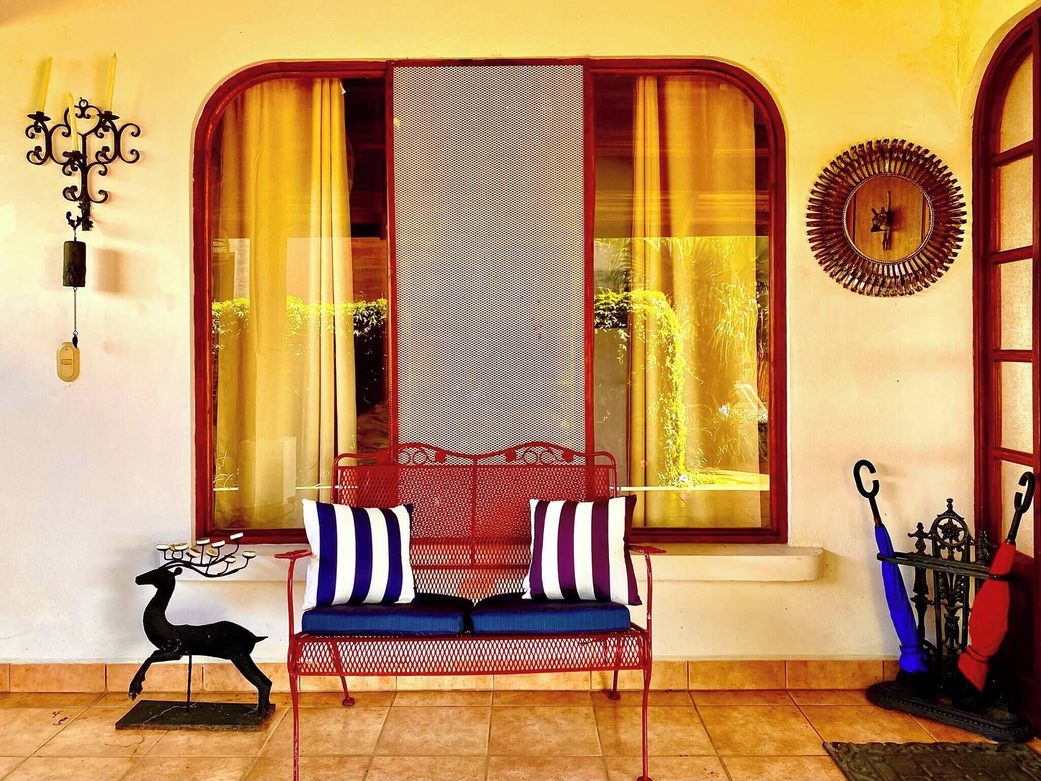 A room with a red metal chair, a blue and white striped cushion, a decorative wall clock, and a window with yellow curtains.