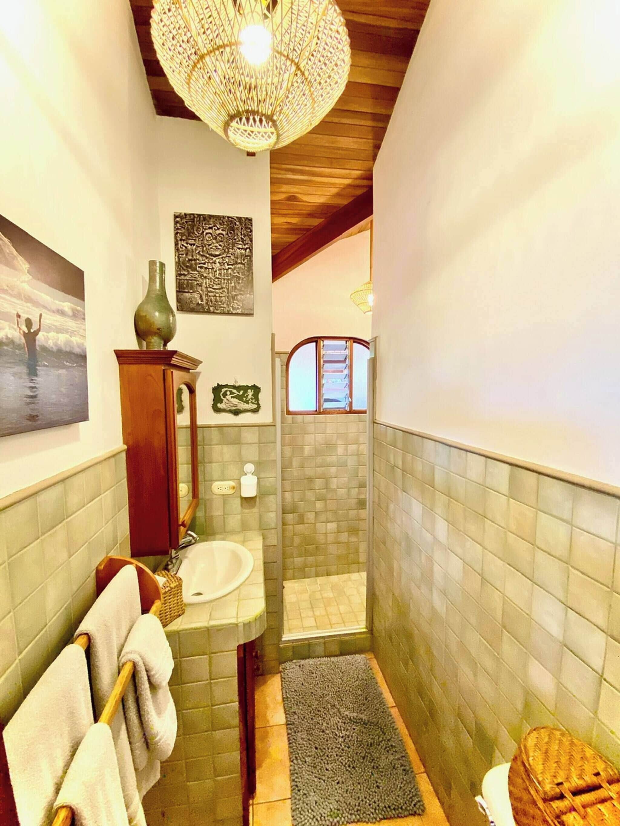 Un baño con tocador de madera, lavamanos, espejo, perchero para toallas, un jarrón verde y una ducha con pared de azulejos.