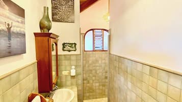 Un baño con tocador de madera, lavamanos, espejo, perchero para toallas, un jarrón verde y una ducha con pared de azulejos.