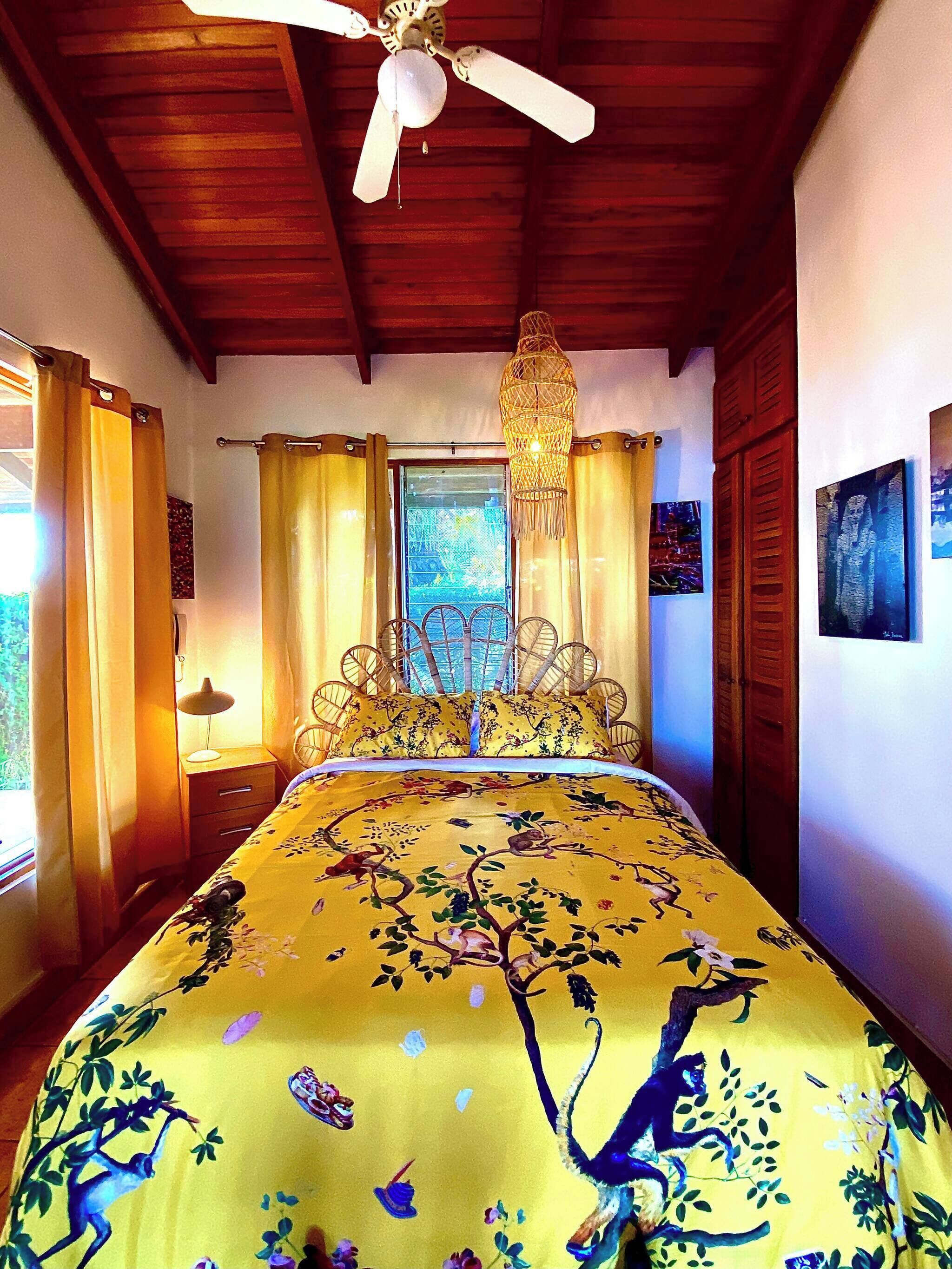 Quarto com cama, ventilador de teto, janela com cortinas e luminária na parede.