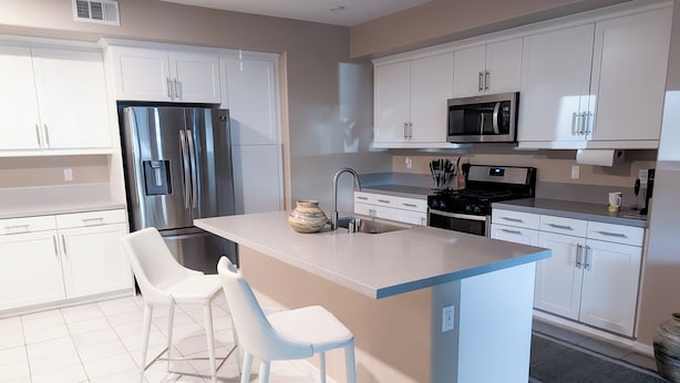 Une cuisine moderne avec des meubles blancs, un réfrigérateur en acier inoxydable, un four à micro-ondes et un îlot central agrémenté de deux chaises blanches.