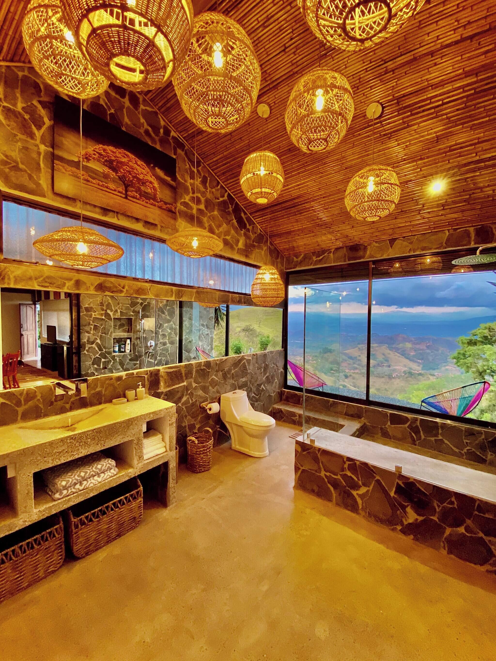 A bathroom with a stone wall, a toilet, a sink, and a large window with a view of the landscape.